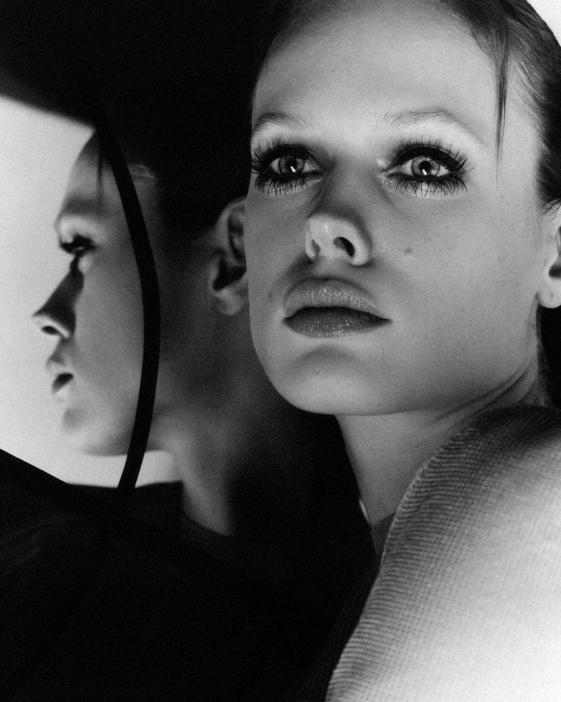Striking black and white studio portrait of a woman with long eyelashes and reflective surface. A face reflects the light.