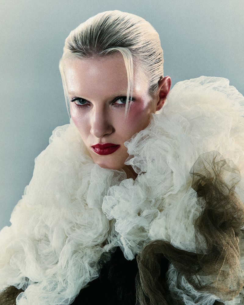 Portrait of a woman with a severe gaze and red lipstick, framed by a large ruffled tulle collar, against a muted blue background.