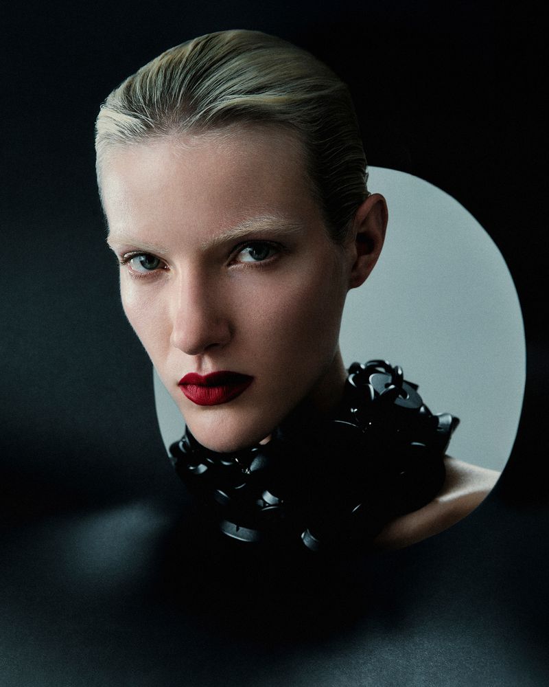Close-up portrait of a woman with fair skin and dark red lipstick, wearing a glossy black necklace against a black and gray gradient backdrop.