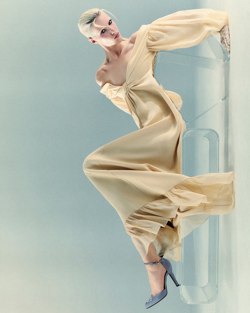 Woman in a beige dress and high heels posing next to a clear acrylic column on a light blue background in a fashion editorial shot.
