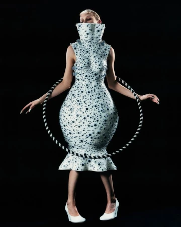 Woman in an avant-garde white dress with black dots posing in a studio. The dress has a high collar and is paired with white heels.