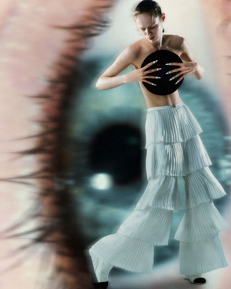 A stylish, slender adult poses with white tiered pants, holding a black disc in front of a giant eye background, fashion editorial.