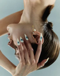 Close-up portrait of a model with long pointed stiletto nails featuring painted eye designs, wearing pearl earrings.