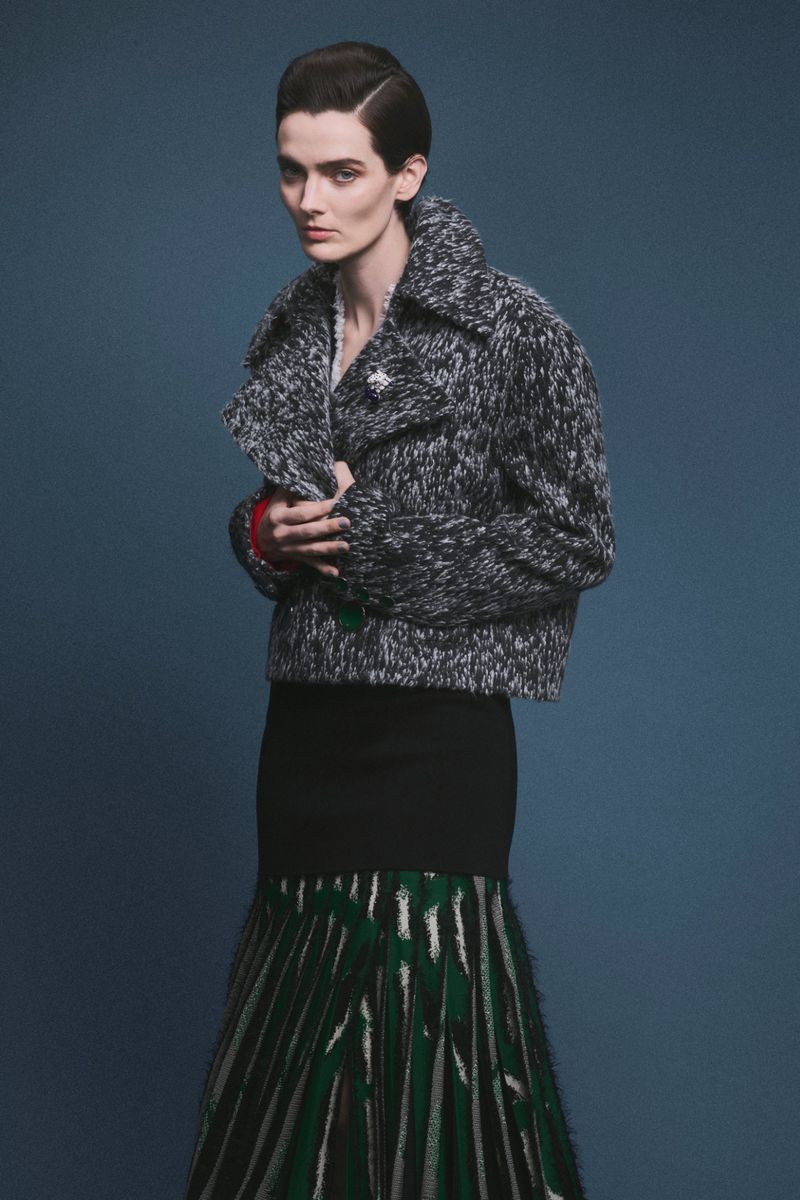 Model wearing a grey tweed jacket and patterned skirt against a dark studio background