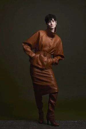 Model with short dark hair wearing a matching brown leather jacket and skirt with knee-high boots against a dark studio background