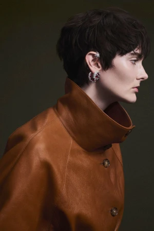 Profile portrait of a model with short dark hair wearing a brown leather coat and sparkling earrings.