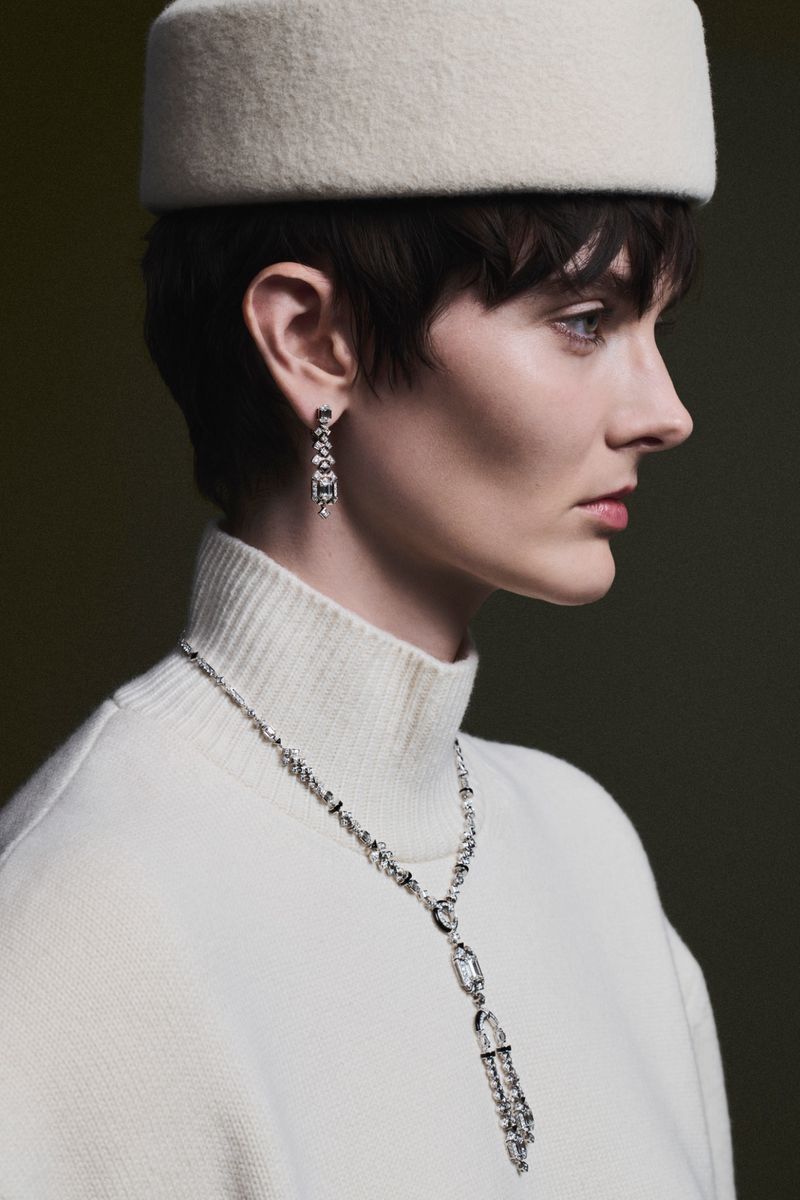 Model in profile wearing a white pillbox hat, white turtleneck, and diamond jewelry against a dark background.
