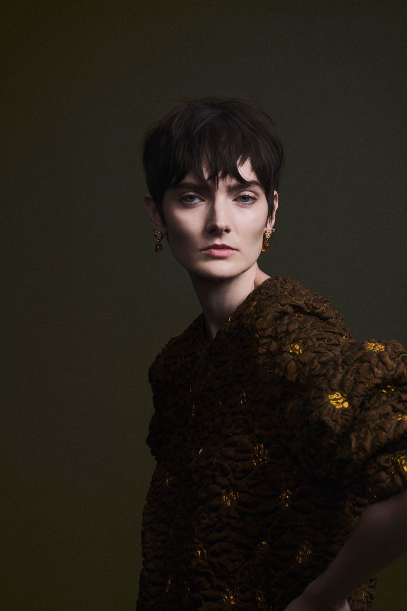 Model with short dark hair wearing a textured brown garment against a dark green backdrop