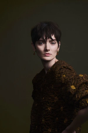 Model with short dark hair wearing a textured brown garment against a dark green backdrop
