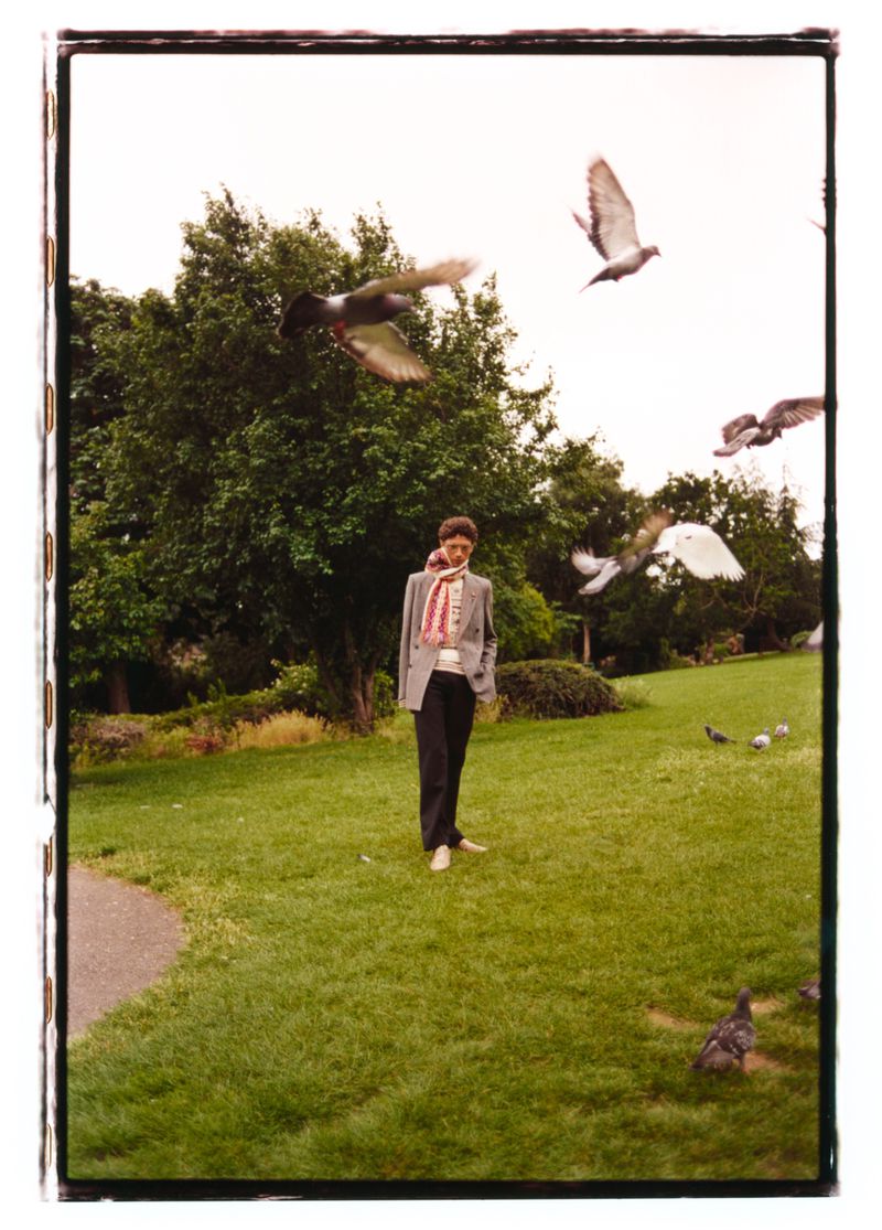 Model standing on grass in a park surrounded by flying pigeons under an overcast sky.