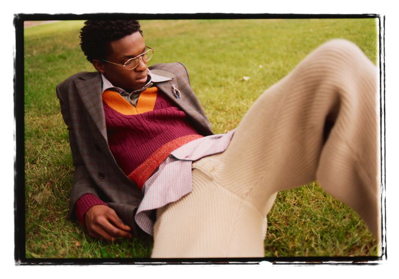 A man wearing glasses, a patterned sweater, and a jacket lying on green grass outdoors.