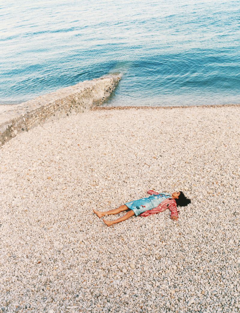Model lying on a pebbled beach beside the ocean, captured from a high-angle view for Chanel Out To Sea editorial.