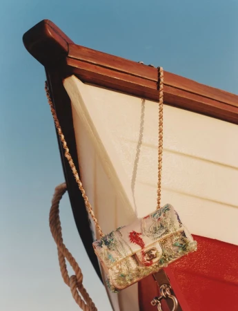 Embroidered flap handbag with interwoven chain strap hanging from the wooden bow of a white and red boat against a clear blue sky.