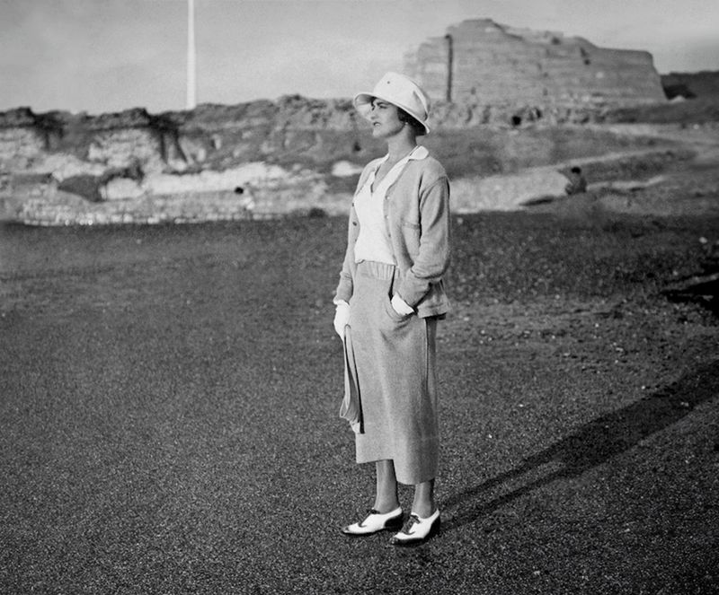 Model standing outdoors wearing a cardigan, skirt, hat, and two-tone shoes against a coastal landscape background
