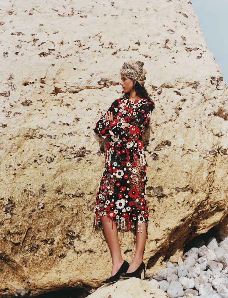 Model standing against a large rock formation wearing a patterned floral dress and matching headwrap with black heels.