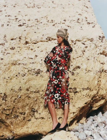 Model standing against a large rock formation wearing a patterned floral dress and matching headwrap with black heels.