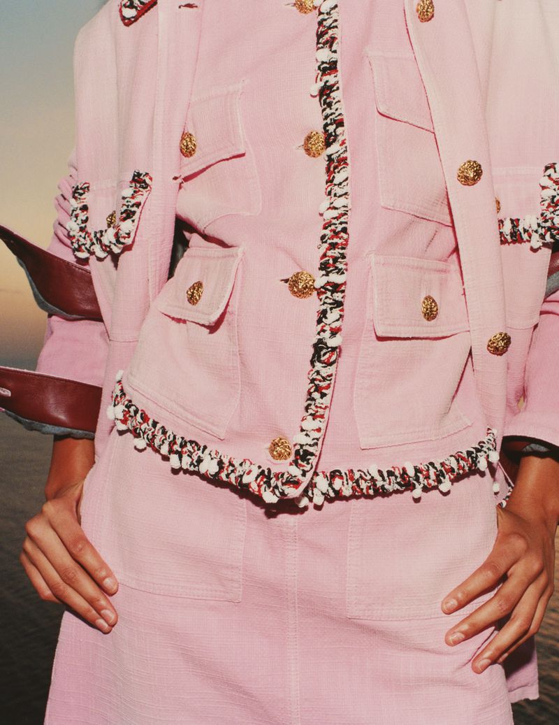 Model wearing a pink tweed jacket and skirt set with ornate gold buttons and fringe trim outdoors by water at sunset
