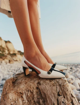 Close-up of a model's legs wearing white and black two-tone heels while standing on a piece of driftwood near a rocky coast.