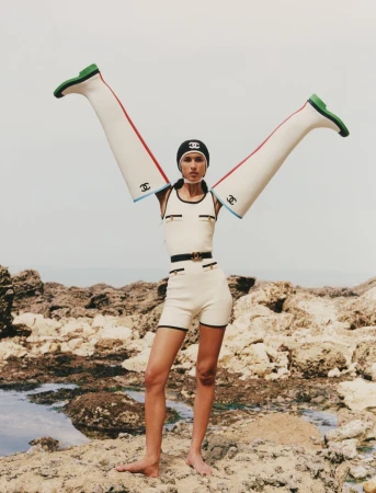 Model standing on a rocky coast wearing a cream romper with tall white boots placed on her outstretched arms