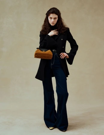 Model in black jacket and dark blue flared jeans holding a brown handbag against a neutral textured studio background.