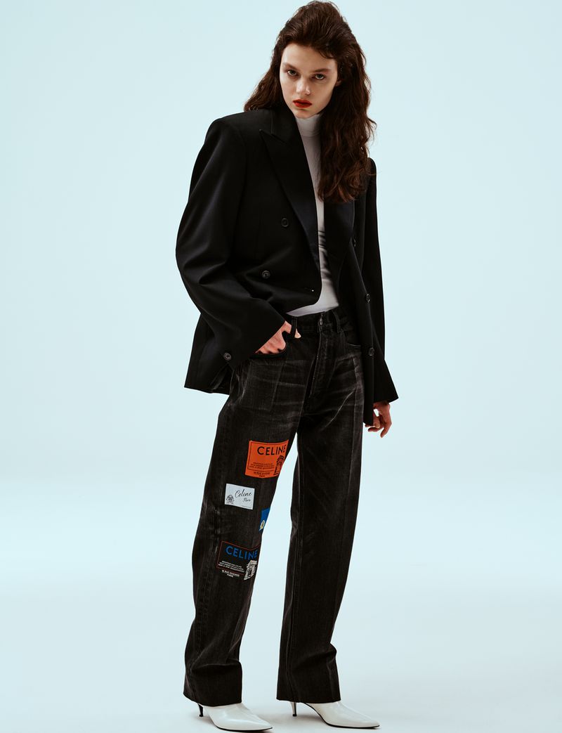 Model in black blazer and patched jeans wearing white heeled boots against a white studio background