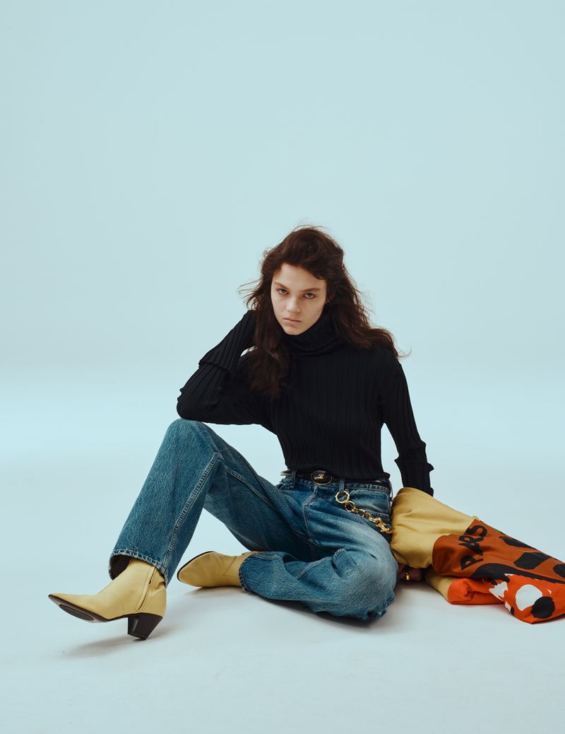 Model seated on a light blue backdrop wearing a black turtleneck, denim jeans, and yellow boots