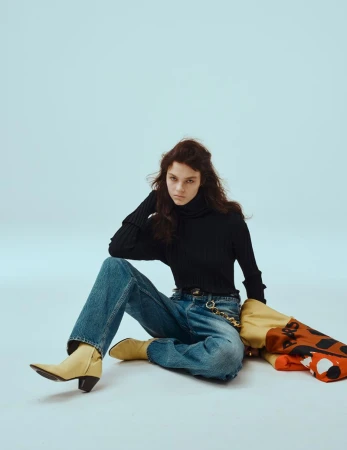 Model seated on a light blue backdrop wearing a black turtleneck, denim jeans, and yellow boots