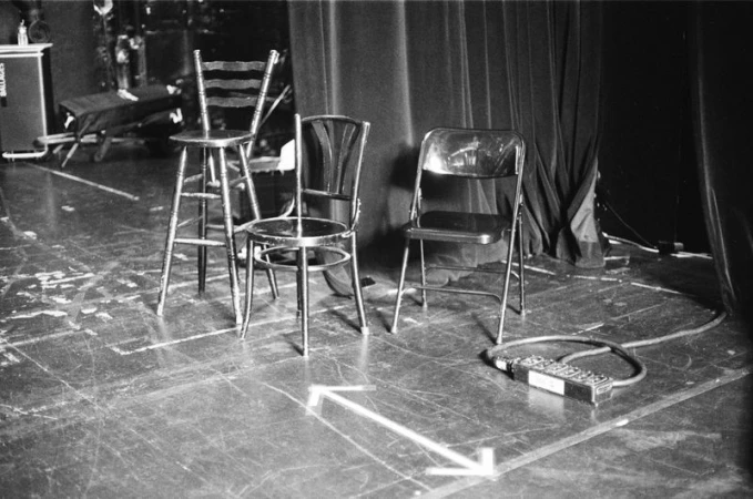 Three different chairs arranged on a wooden stage backstage with dark curtains and a power strip on the floor.