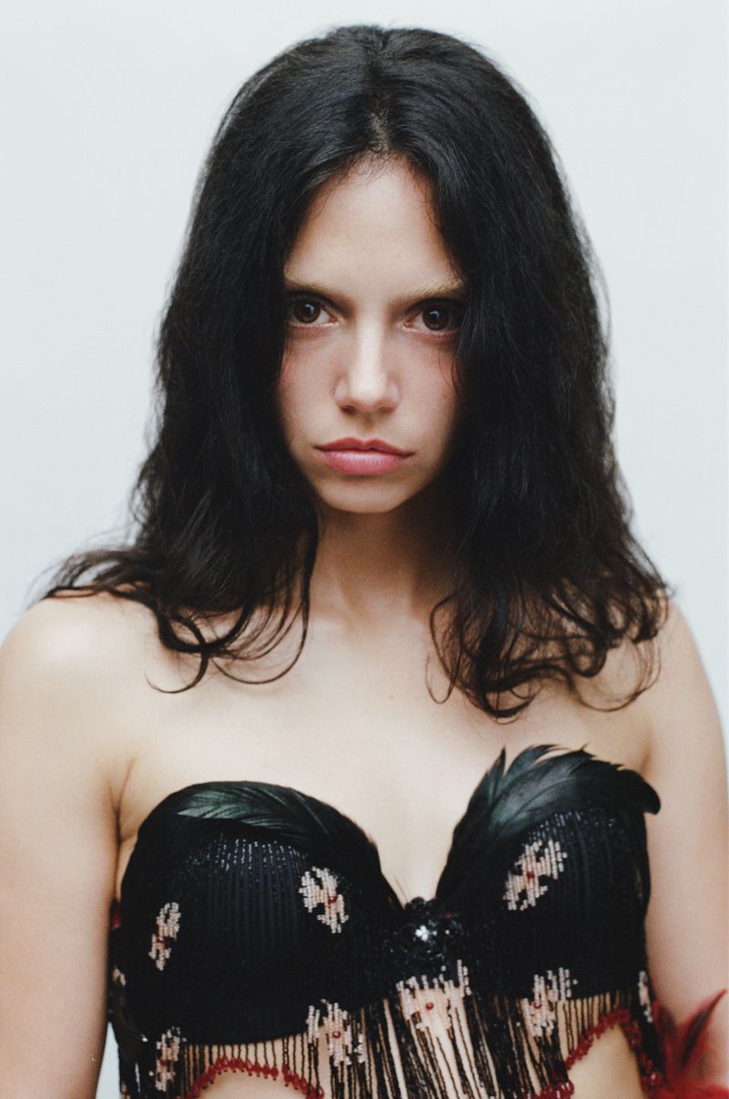 Portrait of a woman with long dark hair wearing a black feather-trimmed strapless top against a white background.