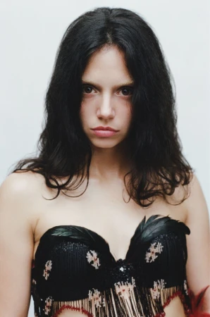 Portrait of a woman with long dark hair wearing a black feather-trimmed strapless top against a white background.
