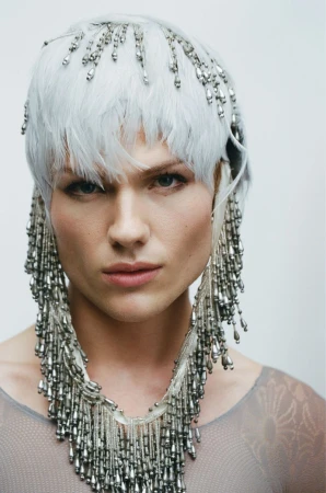 Close-up portrait of a model wearing a white feathered headpiece and elaborate silver beaded necklace against a plain white backdrop.