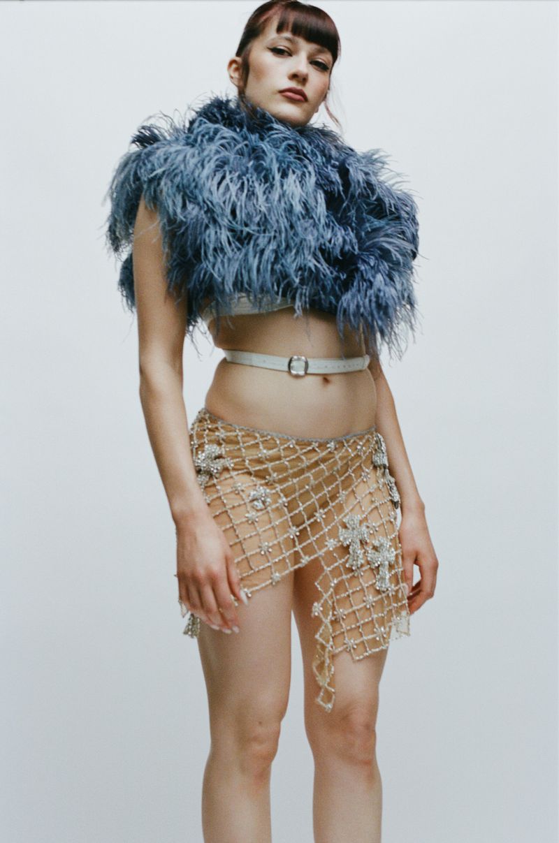 Model wearing a blue feather stole and sheer embellished skirt standing against a plain white studio background.