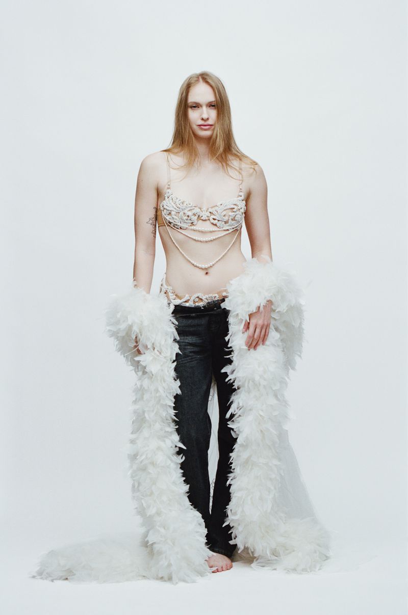 Model wearing embellished bra, black trousers, and white feather boa against a plain white background for Vogue France.