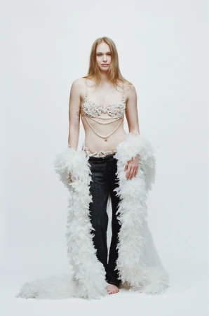 Model wearing embellished bra, black trousers, and white feather boa against a plain white background for Vogue France.