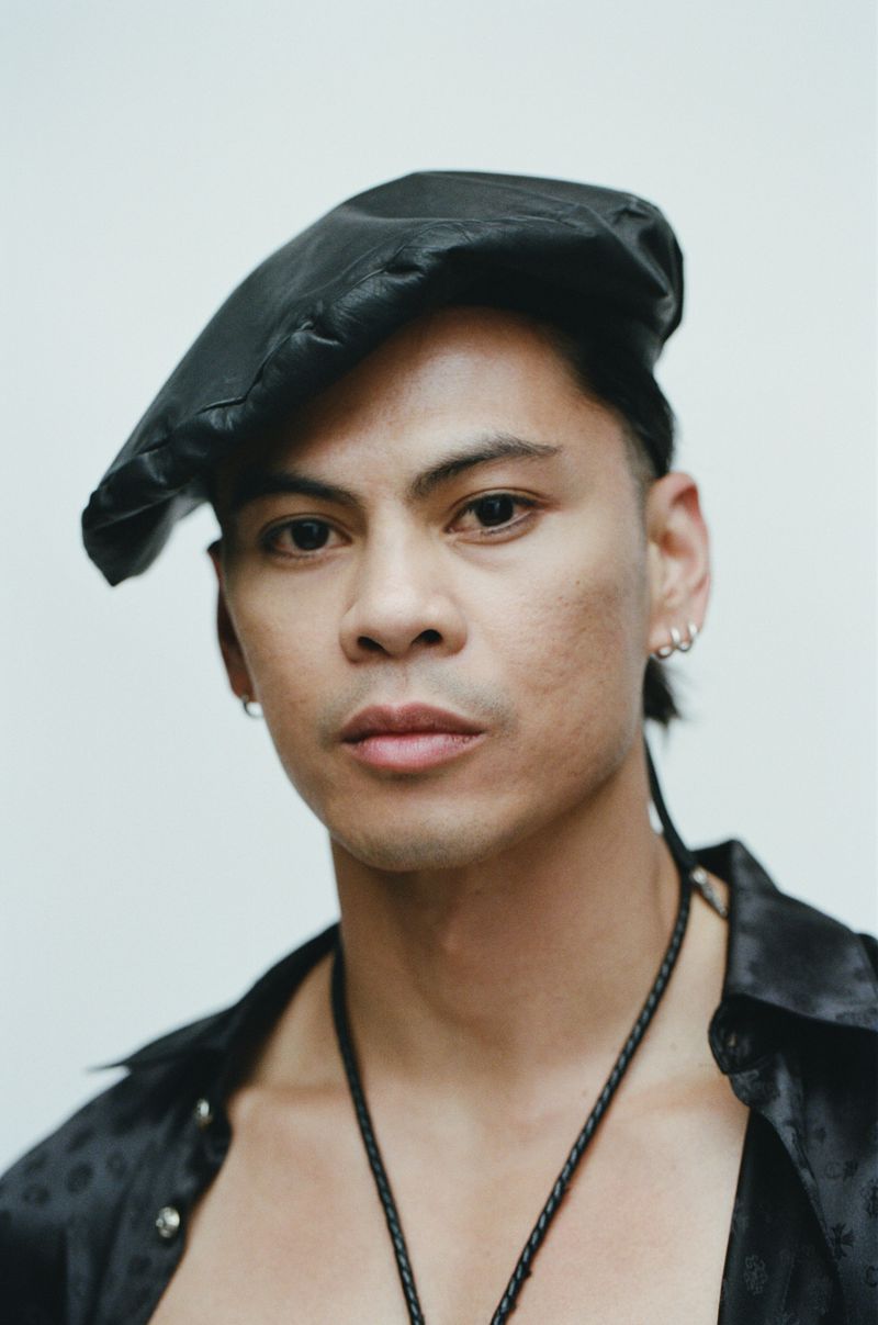 Man wearing a black beret and unbuttoned satin shirt against a white backdrop for Vogue France by Estévez and Belloso.