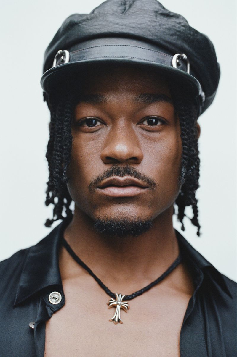 Close-up portrait of a black man wearing a leather cap and necklace against a white background