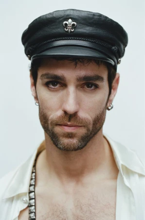 Man wearing a black leather captain hat with a silver emblem and a white shirt against a plain light background