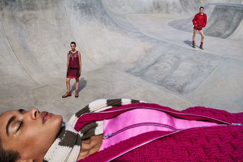 Model lying in foreground wearing magenta knitted zip-up garment with two standing models posed in a large concrete skatepark bowl.