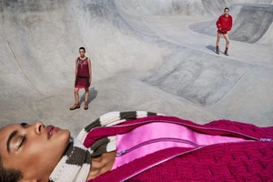 Model lying in foreground wearing magenta knitted zip-up garment with two standing models posed in a large concrete skatepark bowl.