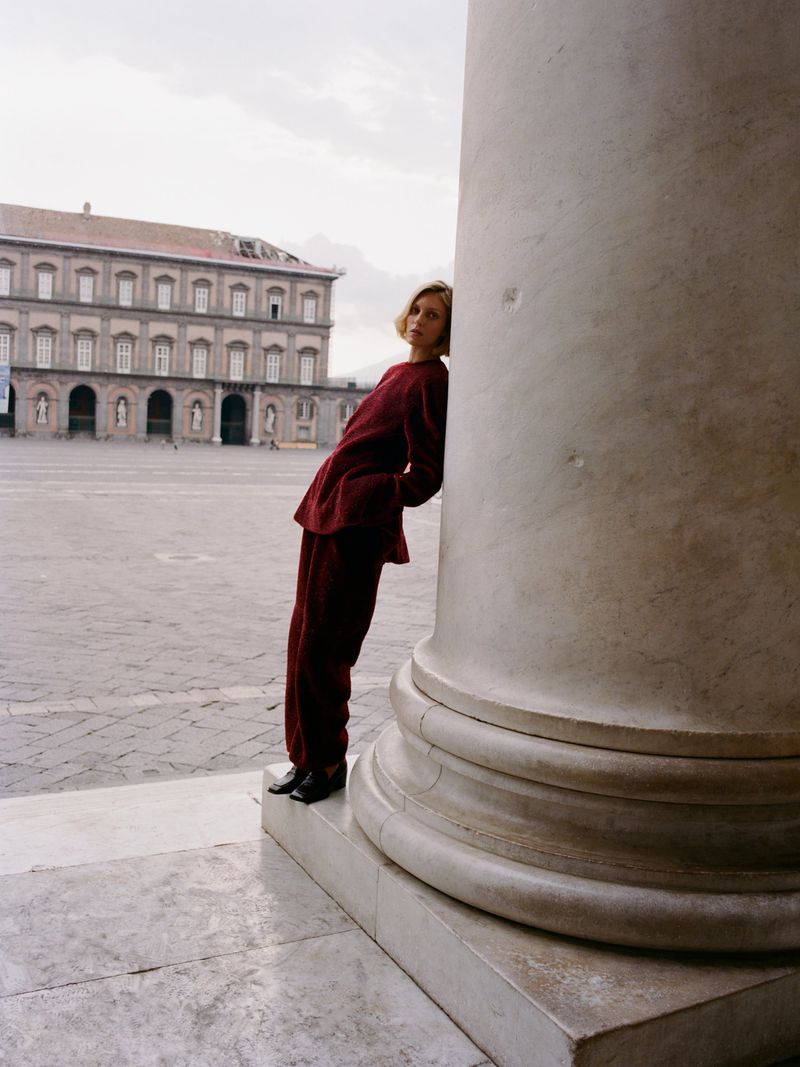 Model leaning against a large classical column in a public square wearing a burgundy top and matching trousers