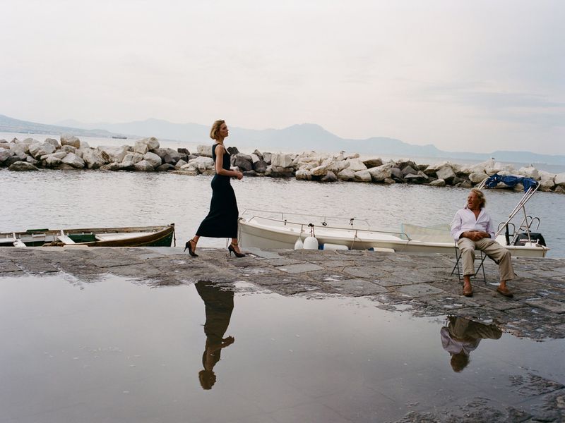 Model Anja Rubik walking along a stone pier by the water with a seated man in the background