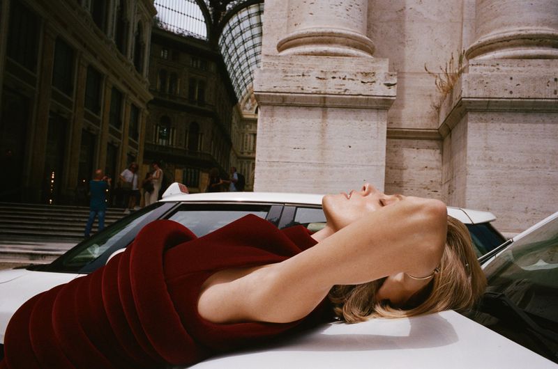 Model Anja Rubik leaning back on a white car hood wearing a structured red dress against an urban architectural street background