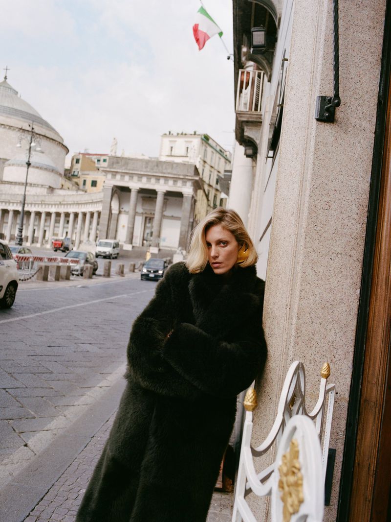 Model Anja Rubik leaning against a stone building on a city street wearing a dark fur coat.