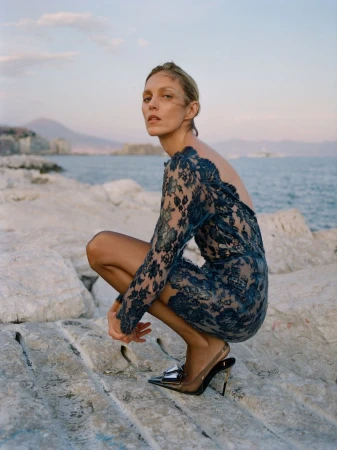 Model Anja Rubik crouching on rocky shore wearing blue lace dress for Financial Times HTSI Magazine