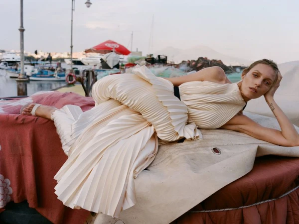 Model Anja Rubik lying on a boat at a harbor wearing a pleated cream dress photographed by Max Farago
