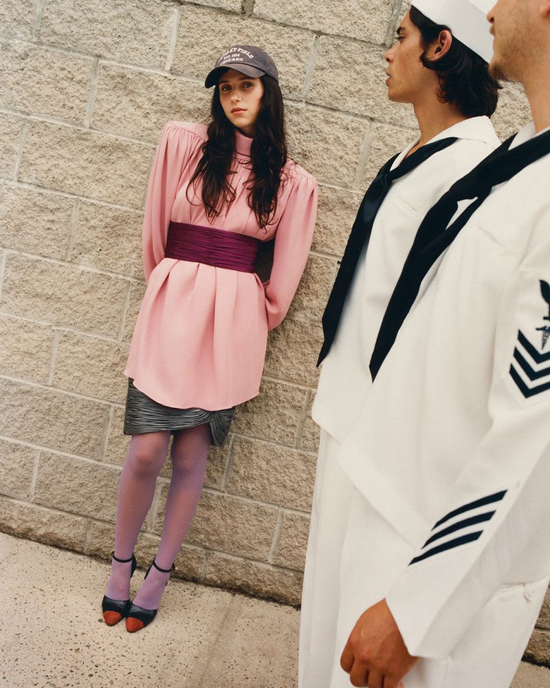 Woman in pink dress and cap leaning against a brick wall near two men in white sailor uniforms