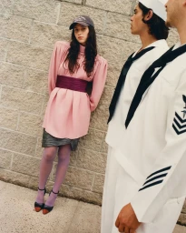 Woman in pink dress and cap leaning against a brick wall near two men in white sailor uniforms