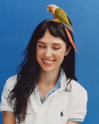 Portrait of Clairo with a green and red parrot perched on her head against a solid blue background