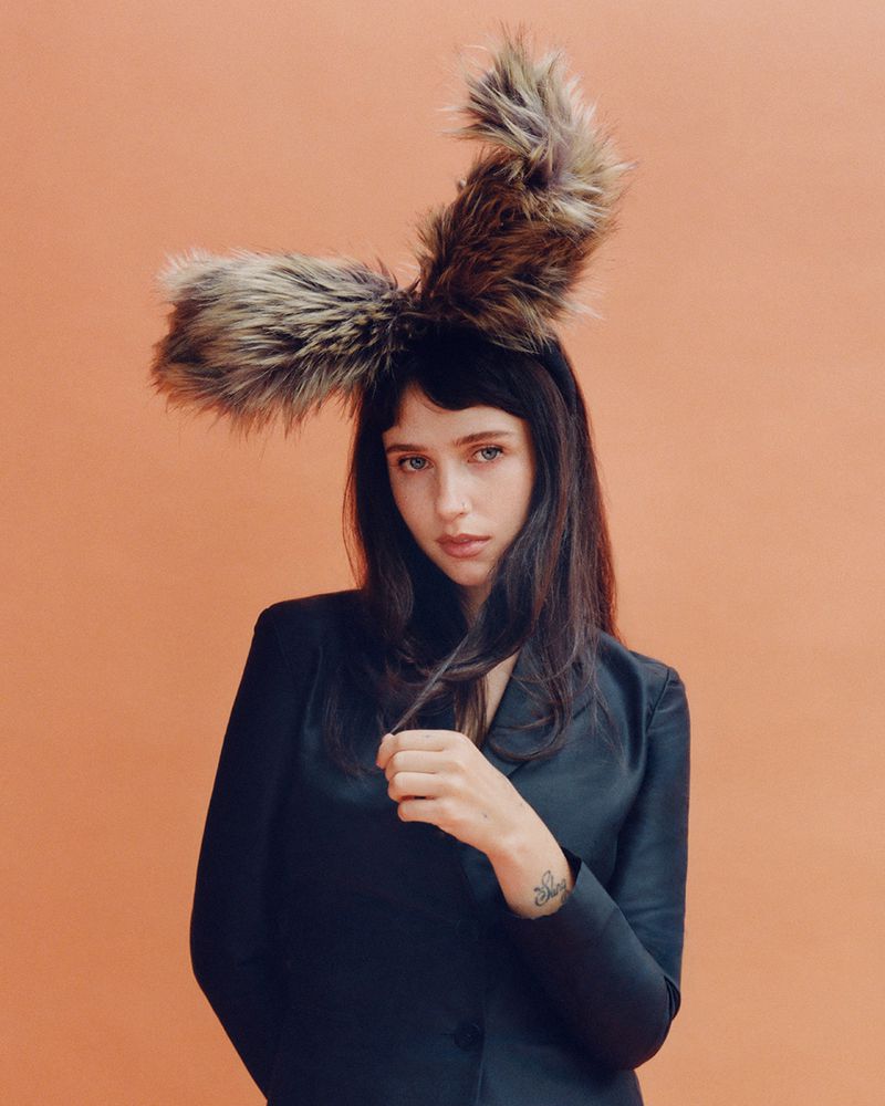 Clairo wearing a dark blazer and large faux-fur animal ear headband against an orange studio background