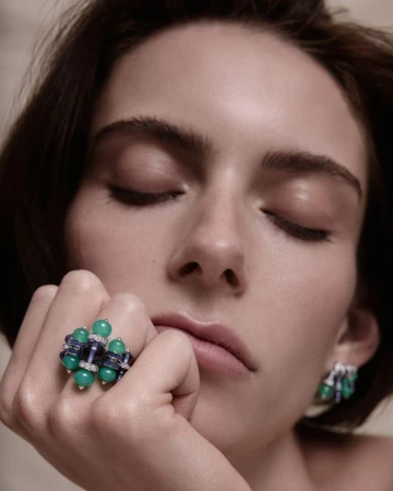 Close-up portrait of a model wearing ornate green and purple beaded rings and matching earrings against a neutral studio background.